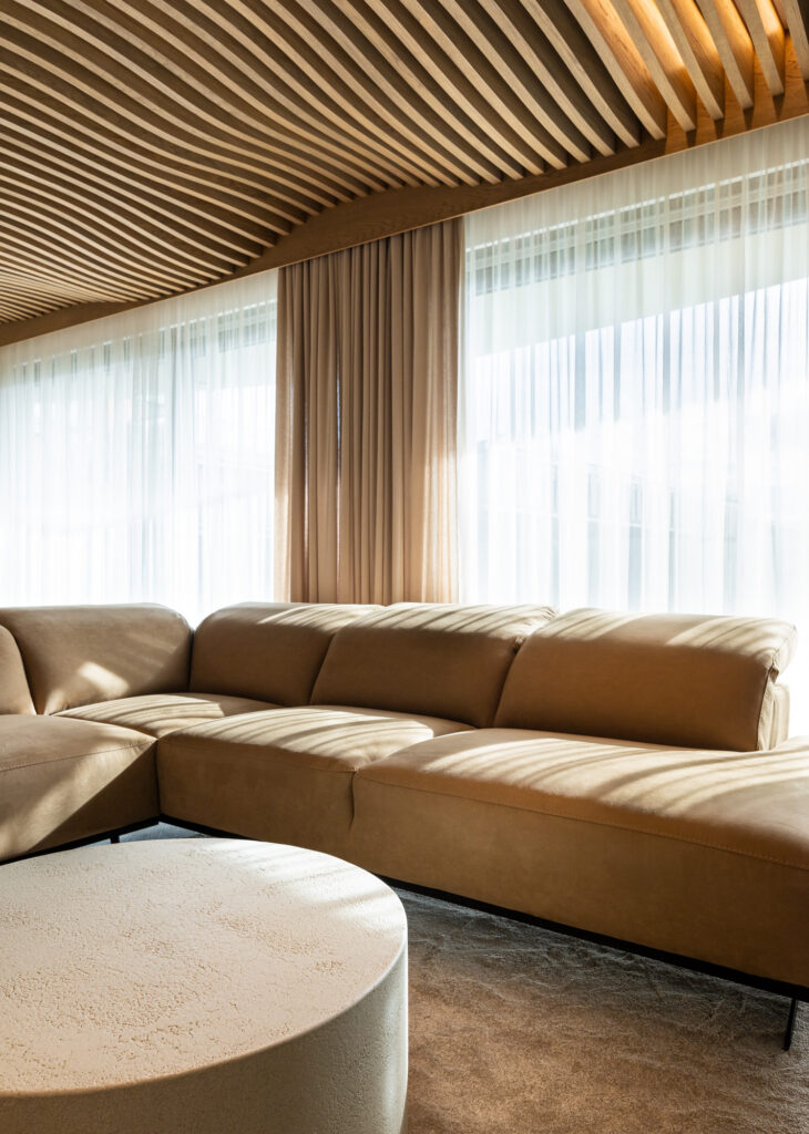 Airwood wave-form wooden lamella ceiling above a beige sofa, with sheer curtains in a luxury apartment