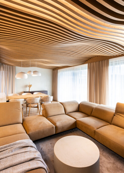 Living-room corner with a beige sofa, the Airwood wooden lamella ceiling flowing overhead by the windows