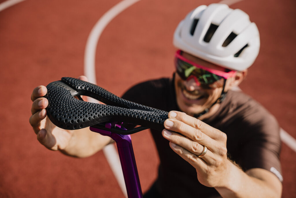 Smiling cyclist in a helmet and sunglasses holding up a black 3D-printed Posedla Joyseat saddle to the camera