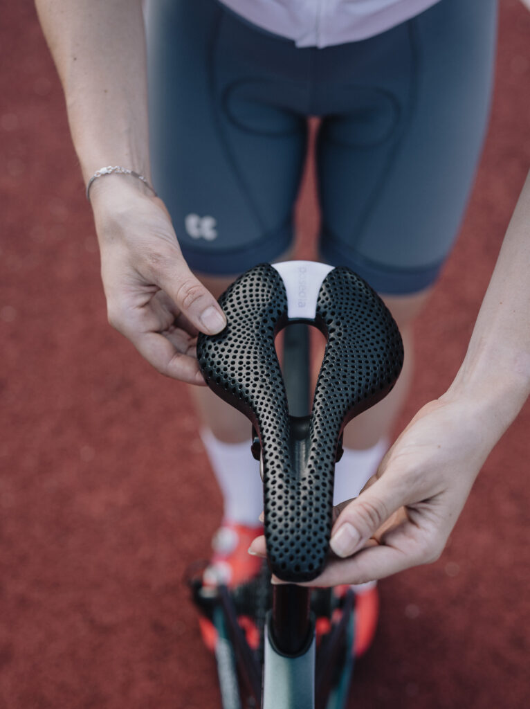 Cyclist holding a black 3D-printed Posedla Joyseat saddle above the seatpost with both hands, viewed from above