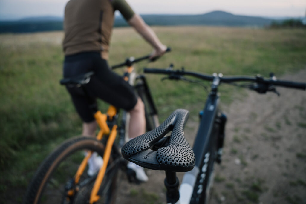 Black 3D-printed Posedla Joyseat saddle in focus on a bike, two riders on a hilltop at dusk behind