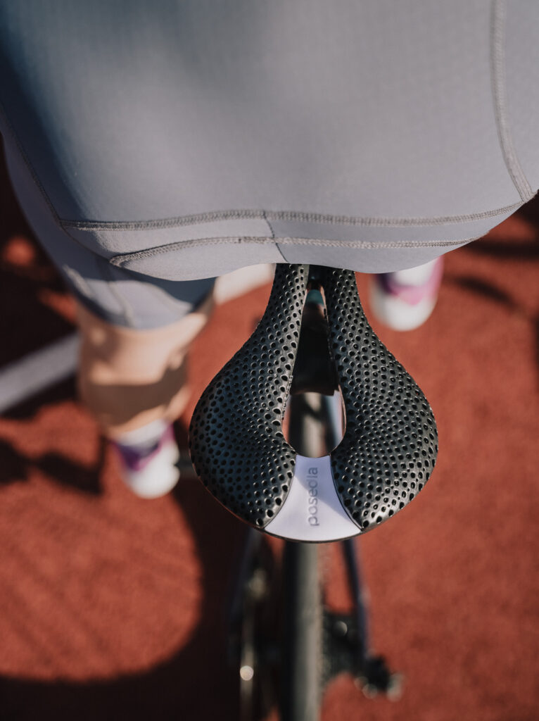 Top-down over-the-shoulder view of a black 3D-printed Posedla Joyseat saddle beneath a cyclist in grey kit